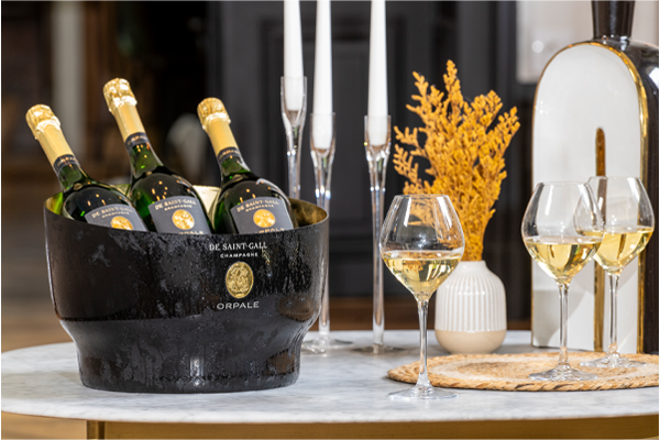 Drei Flaschen De Saint-Gall Orpale Champagner im schwarzen Kühler, daneben drei gefüllte Gläser auf Marmortisch.
