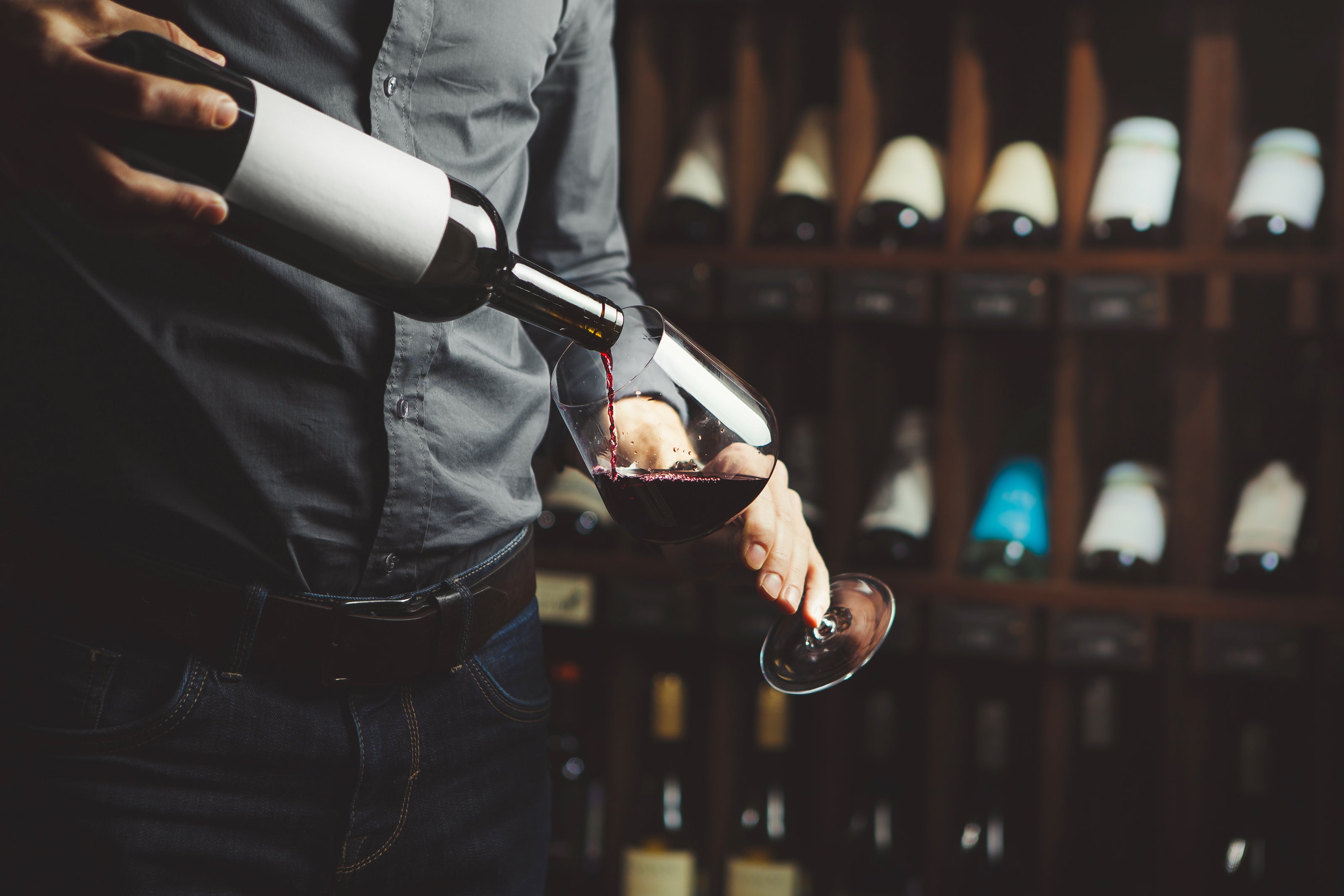 Mann schenkt Rotwein in ein Weinglas ein, im Hintergrund ein Weinkeller.